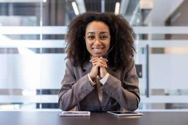 Ofiste oturan takım elbiseli gülümseyen Afro-Amerikan bir kadının portresi. Kulaklık takmış, elleri kavuşturulmuş ve kameraya bakıyordu..