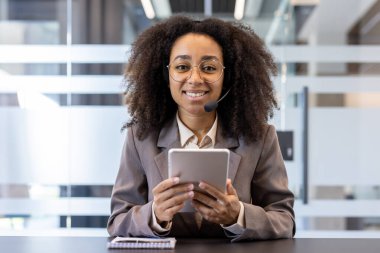 Takım elbiseli ve kulaklıklı gülümseyen Afro-Amerikan bir kadının portresi. Kameraya gülümsüyor, masada oturuyor ve elinde tablet tutuyor..