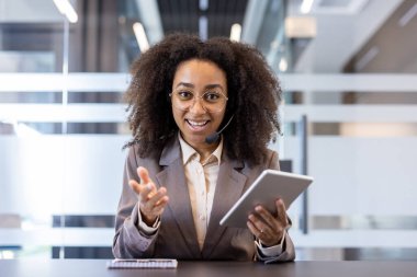 Genç Afro-Amerikan bir kadının portresi, takım elbiseli, kameranın önünde bir masada oturuyor, elinde bir defter tutuyor, kameraya konuşuyor ve el kol hareketi yapıyor..