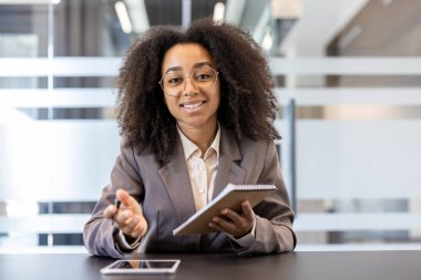 Genç Afro-Amerikalı iş kadını, ofiste kameranın önünde oturuyor ve konuşuyor, antrenman yapıyor, elinde kalem ve defter tutuyor..
