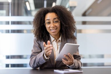 Takım elbiseli ve gözlüklü gülümseyen Afro-Amerikan bir kadın ofiste bir masada oturuyor, elinde bir not defteri ve kamerayla konuşuyor..