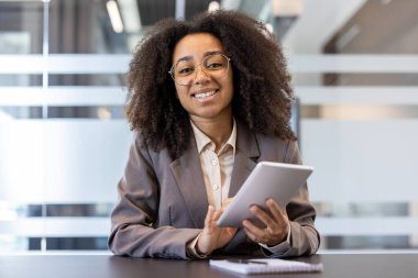 Ofisteki bir masada oturan, tablet kullanan ve gülümseyerek kameraya bakan takım elbiseli Afro-Amerikan bir kadının portresi..