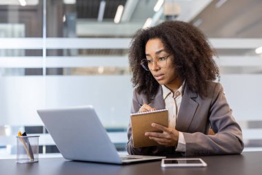 Konsantre olmuş, takım elbiseli Afro-Amerikan kadın ofiste dizüstü bilgisayarın önünde oturuyor ve notlar alıyor..