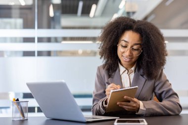 Konsantre olmuş ve gülümseyen genç Afro-Amerikan kadın elinde dizüstü bilgisayarla büroda oturuyor ve bir not defterine notlar alıyor, bir konferansı dinliyor ve uzaktan ders çalışıyor..