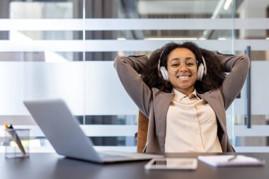 Sonuçtan memnun bir şekilde gülümseyen bir gencin portresi ve işyerindeki bir sandalyede rahatça oturan kulaklıklı Afrikalı Amerikalı bir iş kadınının işi..