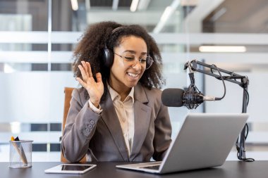 Gülümseyen, takım elbiseli, mikrofonun önünde kulaklık takan ve bilgisayar başında konuşan, selamlayan ve ekranda el sallayan genç Afrikalı Amerikalı kadın..