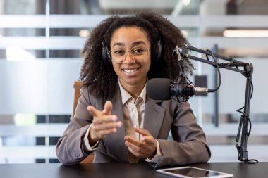 Takım elbiseli genç Afrikalı Amerikalı bir kadının portresi. Masada kulaklık takıyor ve mikrofonlu bir standın arkasında kameraya doğru konuşuyor..