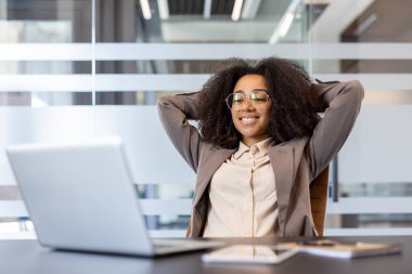 Gülümseyen ve tatmin olmuş genç bir Afro-Amerikan kadın masasında oturuyor gözleri kapalı ve ellerini sırtına koyup dinleniyor..