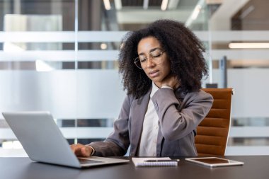 Yorgun bir Afrikalı Amerikalı genç iş kadını bir ofis masasında oturur, eliyle boynunu ovuşturur, ağrı için masaj yapar..