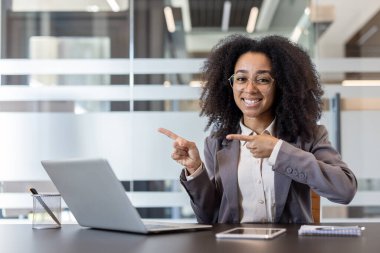 Ofisteki bir masada oturan ve parmaklarıyla boş bir yeri gösteren takım elbiseli gülümseyen Afrikalı Amerikalı bir kadının portresi..