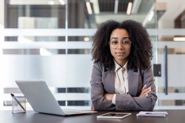 Ciddi ve kendinden emin, takım elbiseli bir Afro-Amerikan kadının portresi bir masada dizüstü bilgisayarla oturuyor ve kameraya bakıyor..