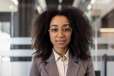 Modern ofiste duran kıvırcık saçlı, gözlüklü, kendine güvenen bir iş kadını. Profesyonel giysiler idari mevcudiyeti vurguluyor. Sakin, sakin ifade zekayı iletir.