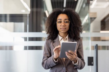 Ofiste elinde tabletle kameraya bakan takım elbiseli ciddi ve kendinden emin bir Afro-Amerikan iş kadınının portresi..