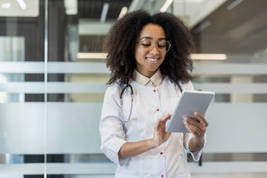 Doktor beyaz önlük giyiyor ve steteskop kullanıyor. Dijital iletişim için tablet kullanıyor. Teknoloji ve sağlık verimliliği üzerine odaklanmış profesyonel bir ortam. Pozitifliğin ifadesi