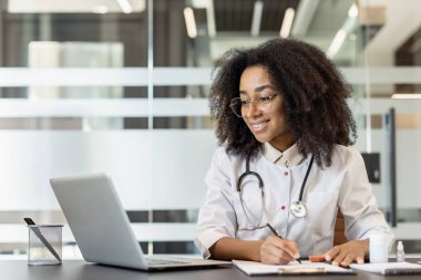 Bilgisayarda çalışan gözlüklü bir doktor modern ofiste kalemle notlar yazıyor. Profesyonel steteskop ve evraklar masanın üzerinde. Çevrimiçi görevlerde bulundu, teknoloji sağlık uzmanlığını birleştirdi.