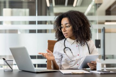 Yoğunlaşmış ve ciddi bir Afro-Amerikalı kadın doktor. Klinikte dizüstü bilgisayarın önündeki masada oturuyor. Elinde tabletle hastaya danışmanlık yapıyor ve video konferansı düzenliyor..