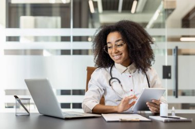 Ofiste bilgisayarla ve tabletle çalışırken gülümseyen kadın doktor. Teknolojinin ve iletişimin verimli bir şekilde kullanıldığını gösteren tıbbi alanda uzman. Sağlık hizmetleri yeniliği kavramı