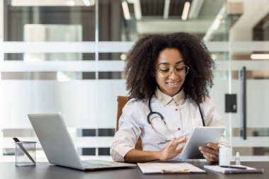 Gülümseyen ve meşgul genç Afrikalı Amerikalı kadın doktor hastane ofisinde bir masada oturuyor ve tablet kullanıyor..