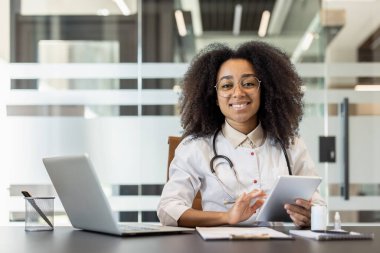 Klinikte beyaz önlüklü, elinde tabletle oturmuş kameraya bakan gülümseyen Afro-Amerikan bir kadının portresi..