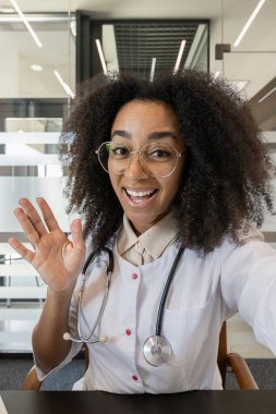 Modern ofisteki video görüşmesi sırasında gözlüklü ve beyaz önlüklü gülümseyen doktor. Teletıp ile ilgilenen dost canlısı sağlık uzmanı, teknoloji hastalarla olan bağlantıyı vurguluyor.