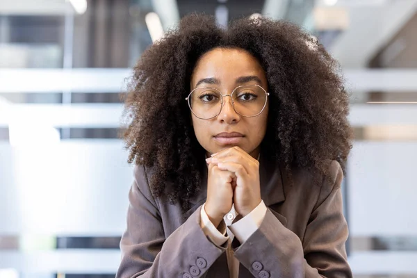 Takım elbiseli ve gözlüklü genç bir Afro-Amerikan kadının yakın plan portresi. Ofisin içinde oturuyor, ellerini birarada tutuyor ve kameraya ciddi ciddi bakıyor..