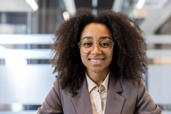 Ofiste takım elbiseli, gülümseyen kıvırcık saçlı, Afro-Amerikan bir kadının kameraya kendinden emin bir şekilde bakarken çekilmiş yakın bir portresi..