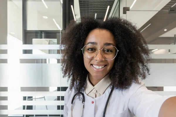 Ofiste selfie çekerken gülümseyen sağlık uzmanı. Gözlük ve steteskop takan kişi, profesyonelliği ve arkadaşlığı iletiyor. Akıllı telefon ile bağlantının görüntüsü alınıyor.