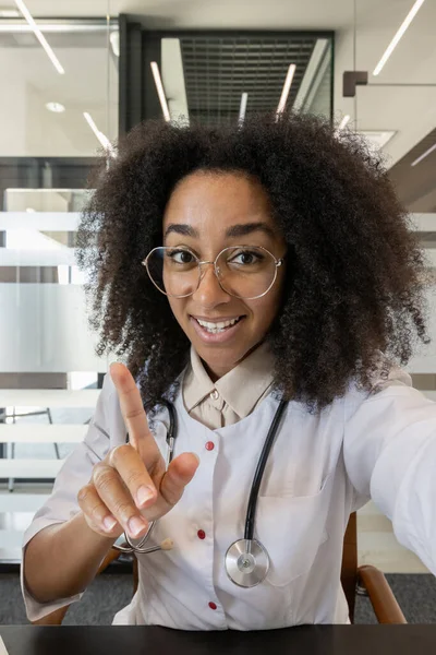 Hastanede genç bir Afro-Amerikalı kadın doktorun dikey yakın plan fotoğrafı. Masada oturuyor ve online video konsültasyonu yapıyor..