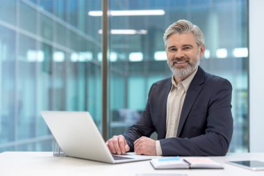 Modern bir ofis masasında sakallı, laptopta çalışırken kameraya güvenle gülümseyen, profesyonel başarıyı ve liderliği somutlaştıran olgun, gri saçlı bir iş adamı.