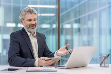 Olgun iş adamı gülümsüyor, el kol hareketi yapıyor ve bilgisayarındaki bir video konferansında not alıyor. Modern ofis ortamında büyük cam pencerelerle çalışıyor.
