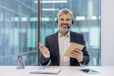 Kulaklık takmış, kameraya gülümseyen, not defteri tutan ve el kol hareketi yapan olgun bir çağrı merkezi ajanı modern ofis ortamında dost canlısı profesyonel müşteri desteği sağlıyor.