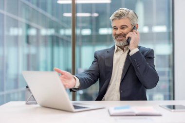 Üst düzey işadamı bir telefon görüşmesi sırasında sırıtarak bilgisayar ekranına el kol hareketi yaparak kendinden emin, profesyonel bir uzaktan çalışma ortamı sunuyor.