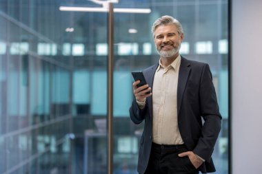 Kendine güvenen, sakallı, gri saçlı, cep telefonu kullanan, modern bir ofis binasında tek eli cebinde duran olgun bir iş adamı.