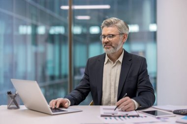 Masada çalışan deneyimli profesyonel işadamı, dizüstü bilgisayarda yazan ve modern bir ofis ortamında çizelgeleri ve raporları inceleyen.