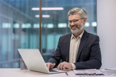 Kıdemli işadamı gülümsüyor, iletişim kuruyor ve ofis belgeleriyle çevrili bir masada iş yapıyor ve modern mimariyi yansıtıyor.
