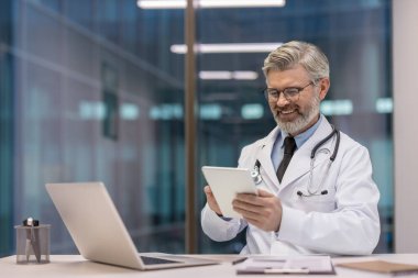 Beyaz önlüklü ve steteskoplu olgun bir erkek doktor bilgisayarlı bir masadaki hasta kayıtlarını incelerken gülümsüyor.