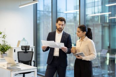 İki farklı iş adamı, parlak çağdaş ofis ortamında bir proje üzerinde işbirliği yaparak önemli belgeler ve finansal verileri tartışıyor.