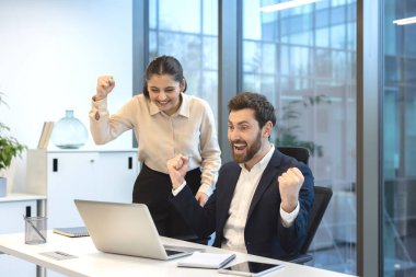 Heyecanlı iş adamları sevinçlerini dile getiriyorlar ve başarıyı kutluyorlar, modern bir ofiste yumruklarını kaldırırken dizüstü bilgisayara bakıyorlar, takım çalışmasını ve başarılı proje tamamlanmasını simgeliyorlar.