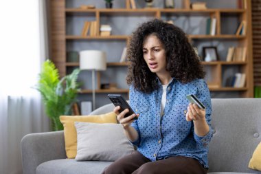 Kanepede oturan genç bir kadın, elinde telefon ve kredi kartı tutarken endişeli ve kafası karışmış görünüyor, evde online alışveriş sırasında ödeme hatasıyla karşılaştı, işlemin başarısız olduğunu vurguladı.