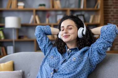 Gözleri kapalı bir kadın, elleri başının arkasında, rahat bir koltukta dinleniyor, kulaklıkla müzik dinliyor, huzur içinde dinleniyor ve evde kendi kendine bakmaktan zevk alıyor.