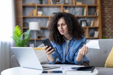 Kıvırcık saçlı kadın akıllı telefonunu ve belgelerini kontrol ediyor. Stres ve kafa karışıklığı yaşıyor. Kişisel mali durumu idare ediyor ve evde bütçe sıkıntısı yaşıyor.