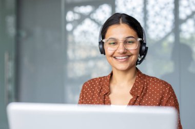Gülümseyen kadın, kulaklığı ve temiz gözlükleriyle, video görüşmesi sırasında dostça uzaktan müşteri desteği sağlıyor, modern ofis ortamında profesyonel ve kendinden emin.