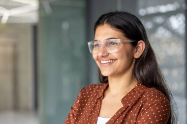 Temiz çerçeveli gözlüklü genç Hintli profesyonel kadın ve kahverengi benekli bluz, modern ofis ortamında dikilirken özgüvenle gülümsüyor, yan görüş vesikalığı
