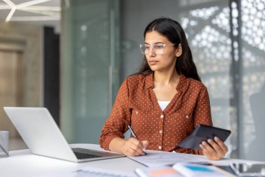 Hintli kadın gözlüklü ve kahverengi gömlekli. Bütçesini hesaplıyor. Elinde hesap makinesi var.