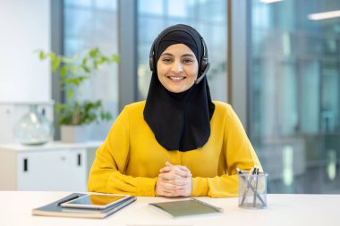 Kendine güvenen Müslüman kadın tesettür ve kulaklık takıyor. Ofis masasında otururken kameraya gülümsüyor. Profesyonel müşteri desteği ve iletişimini gösteriyor.