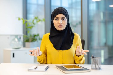 İş kadını, ofis masasında oturan tesettürlü bir kadın, elleri ile hareket ederken kafası karışmış bir ifadeyle doğrudan kameraya bakıyor.