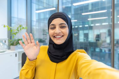 Bir video konferans sırasında tesettür takan, gülümseyen ve kameraya el sallayan Müslüman iş kadını ya da modern bir şirket ofisinde selfie 'ye bakan