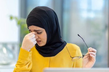 Ofiste çalışırken sürekli başı ağrıyan, gözlerini ovuşturan, gözlük tutan, işle ilgili yorgunluğu ve tükenmişliği tasvir eden Müslüman bir kadın.