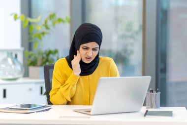 Genç tesettürlü Müslüman iş kadını ofis masasında dizüstü bilgisayarla çalışırken yanağını tutarak diş ağrısı ve stresten konsantre olmaya çalışıyor.
