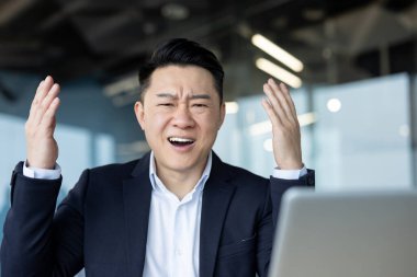 Üzgün bir Asyalı genç adam ofiste dizüstü bilgisayarın önünde oturuyor, hayal kırıklığı içinde ekrana bakıyor, kollarını açıyor..